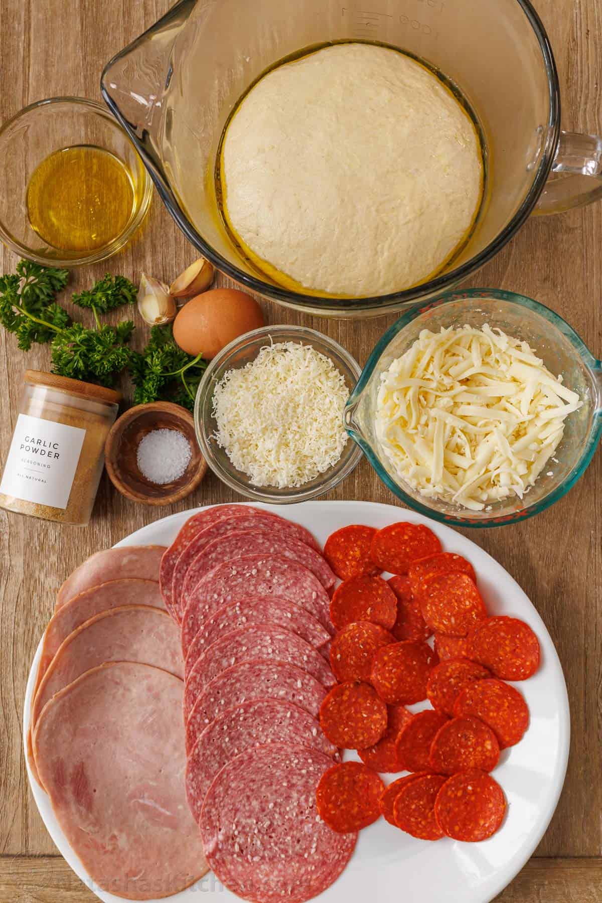 Ingredients for Stromboli with Pizza dough, pepperoni, ham, salami, mozzarella cheese, parmesan cheese, parsley, garlic, salt, garlic powder, and olive oil