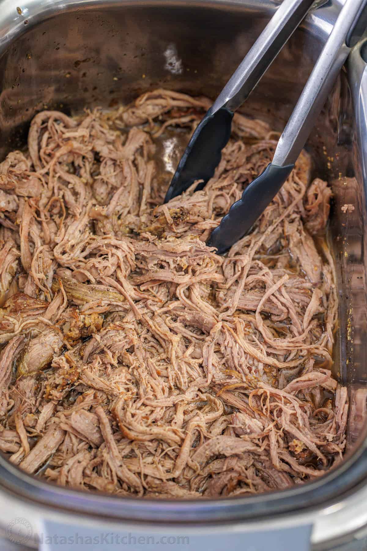 Tongs stuck into shredded beef inside the slow cooker.