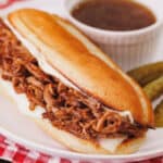 A shredded beef sandwich in a hoagie roll served on white plate next to au jus for dipping.